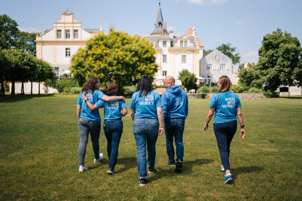 Mitarbeitende der DKB STIFTUNG laufen beim Teamtag 2026 durch den Schlosspark auf Schloss & Gut Liebenberg zu. Sie tragen blaue T-Shirts mit dem Aufdruck „Weil Wir wirkt“.